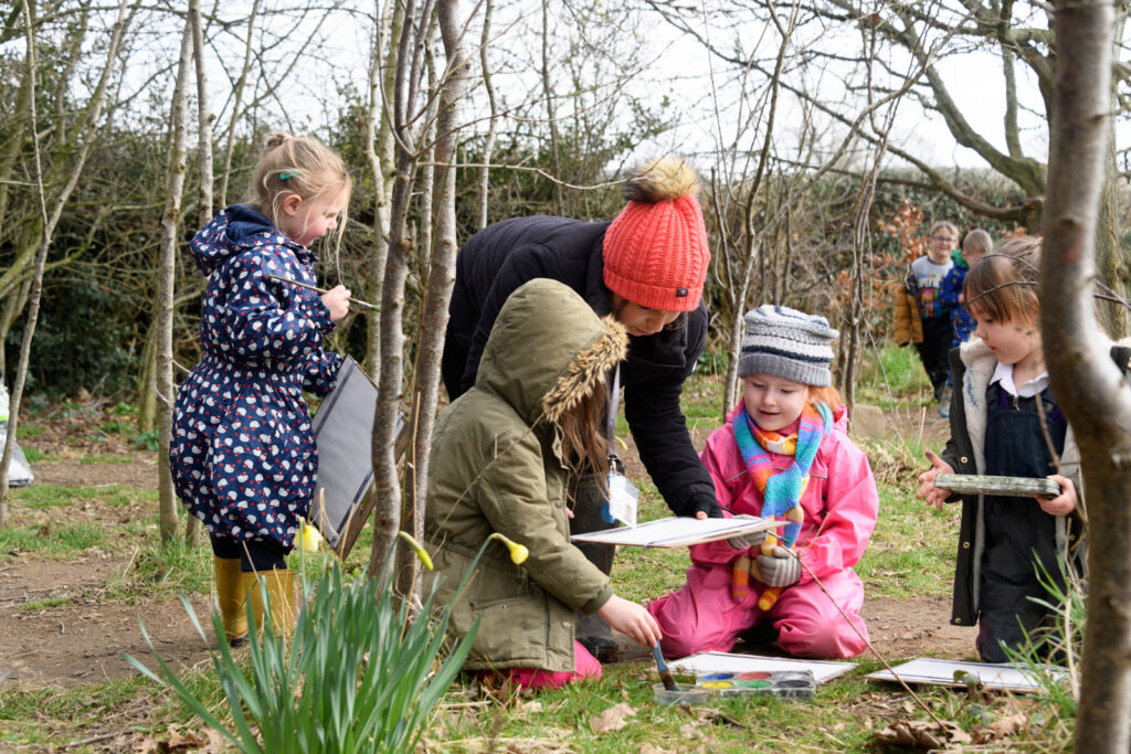 A group of younger students work together using paint to create artwork outside with a teacher helping them.