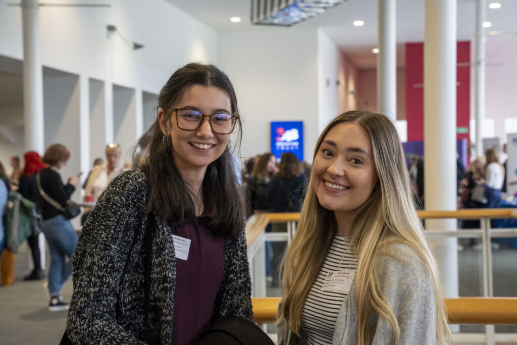 Two people standing indoors at a conference; others are visible in the background.