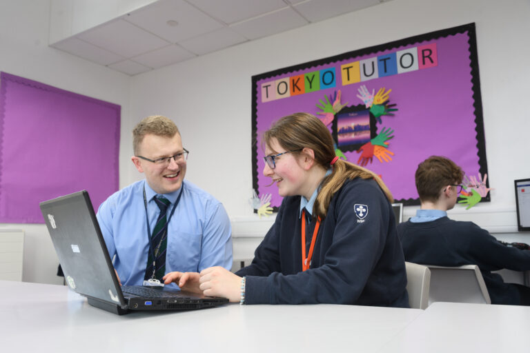 Male teacher and female student sit at a desk with a laptop. Another student and poster display are in the background.