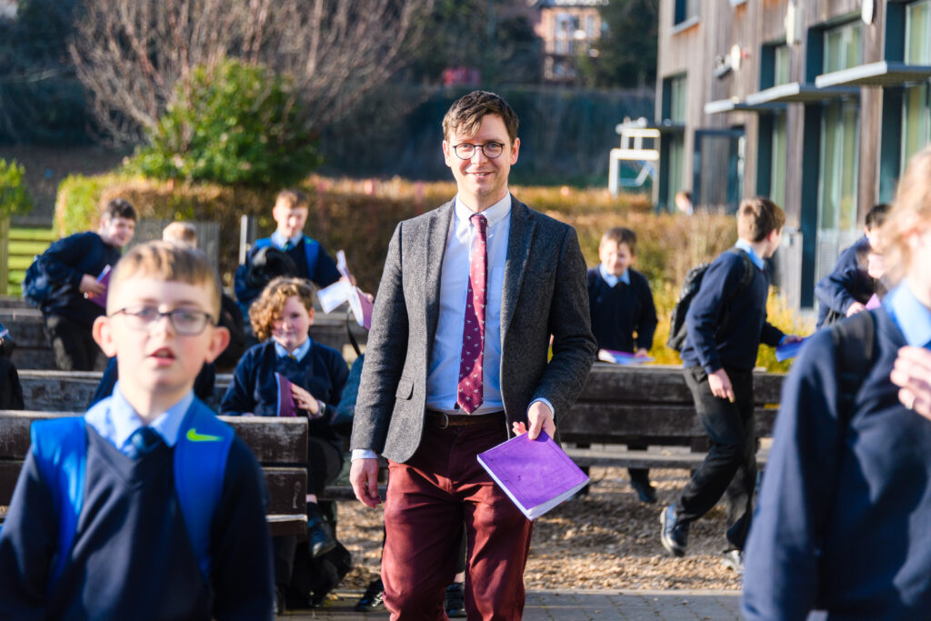 Teacher holding a purple folder walking outdoors toward the camera, with students in uniform sitting or walking nearby.